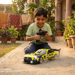 A young boy wearing a light green t-shirt and dark jeans sits on a concrete path in a backyard, smiling while holding a black remote control. In front of him is a bright yellow and black remote-controlled race car toy with 'RACE TEAM' written on the side.