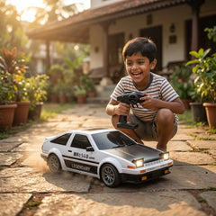 A young boy wearing a striped t-shirt crouches on a stone patio, smiling while holding a black remote control toy car next to a white and black toy sports car.