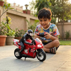 A young boy wearing a colorful graphic t-shirt sits on a paved patio in a backyard, smiling while holding a black remote control. In front of him is a red remote-controlled toy motorcycle with a black rider figure.