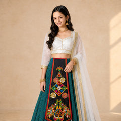 Black | A young girl modeling a traditional Indian ethnic wear set consisting of a white embroidered crop top, a dark teal skirt with a large black embroidered panel, and a sheer white dupatta draped over her shoulder, set against a warm beige background.