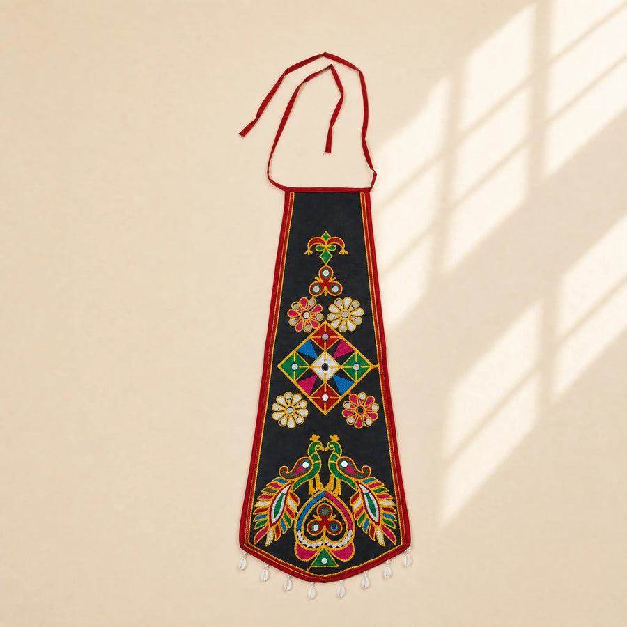 Black | A black embroidered wall hanging or decorative panel with red ties at the top, multicolored floral and geometric motifs, and small white hanging beads along the bottom edge, shown against a warm beige wall with soft window-shadow light.