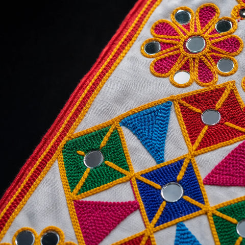 White | Close-up of a white embroidered textile with geometric and floral patterns in red, blue, green, and pink, accented with small round silver mirror embellishments.