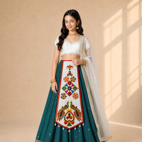 White | A young girl modeling a traditional Indian lehenga choli dress. The outfit features a white embroidered crop top, a teal green skirt with a large white embroidered panel depicting peacocks and floral motifs, and a sheer white dupatta draped over her shoulder.