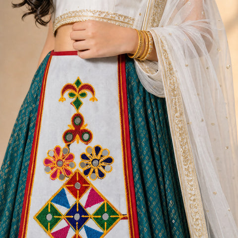 White | Close-up of a traditional Indian outfit featuring a teal blue skirt with a large white embroidered panel and colorful geometric floral motifs, paired with a white dupatta with gold embroidery.