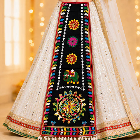 Black | Close-up of a traditional embroidered skirt or lehenga in white with a wide black central panel featuring colorful floral and elephant motifs, plus mirrored embellishments and warm bokeh lights in the background.