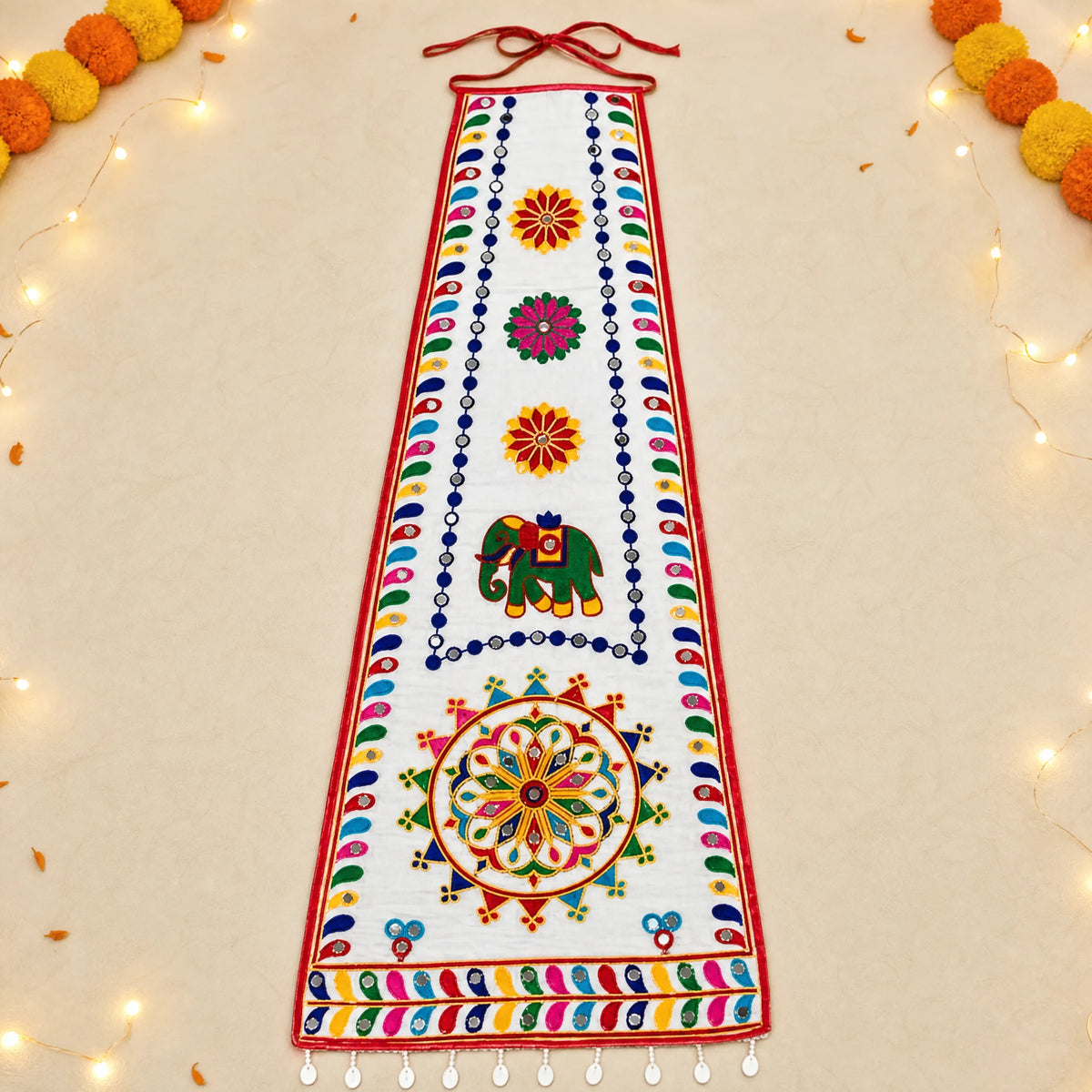 White | A white fabric wall hanging or decorative banner with colorful embroidered patterns, including an elephant motif and floral motifs, displayed flat on a light beige background with decorative string lights and pom-poms around the edges.