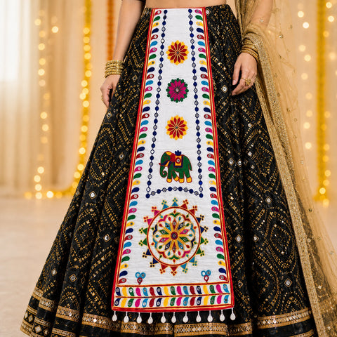White | A close-up of a black and gold embroidered traditional skirt with a large white central panel featuring colorful floral and elephant motifs, shown on a model with a beige dupatta against a warm bokeh background.