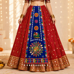 Blue | A red and navy blue embroidered Indian lehenga choli skirt with gold mirror work and colorful motifs, shown on a model in a warm, decorated indoor setting.