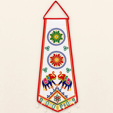 White | A vertical white fabric wall hanging or decorative banner with a red border and hanging loop, decorated with colorful embroidered motifs including circular floral designs, small green clovers, two elephants, and a geometric pattern near the bottom.