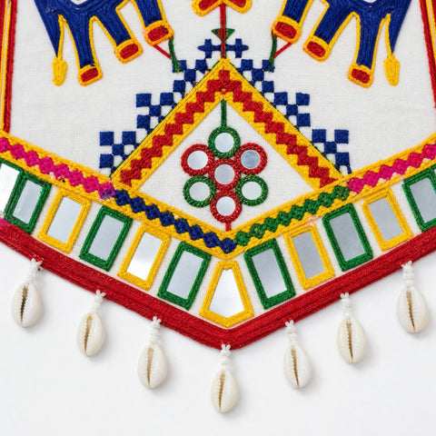 White | Close-up of a white embroidered textile with geometric multicolor stitching, mirrored square details, and hanging cowrie shells along a red border.