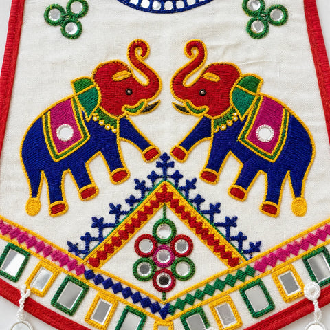 White | Close-up of a white embroidered textile featuring two large blue elephants facing each other, with red trunks and ornate pink, green, yellow, and blue decorative motifs. Mirrored square embellishments and red border trim are visible.