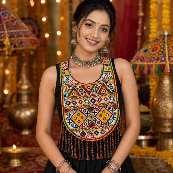 Hira Bharat | A woman wearing a sleeveless black top with an elaborate, colorful geometric embroidered yoke and tassel fringe, posed in a festive, warmly lit indoor setting with decorative lamps and garlands.