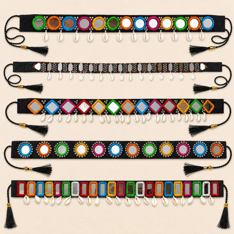 Six decorative waist belts laid flat on a light beige background. Each belt is black with colorful mirrored circular and rectangular embellishments, dangling white cowrie shells, and black tassels at both ends.