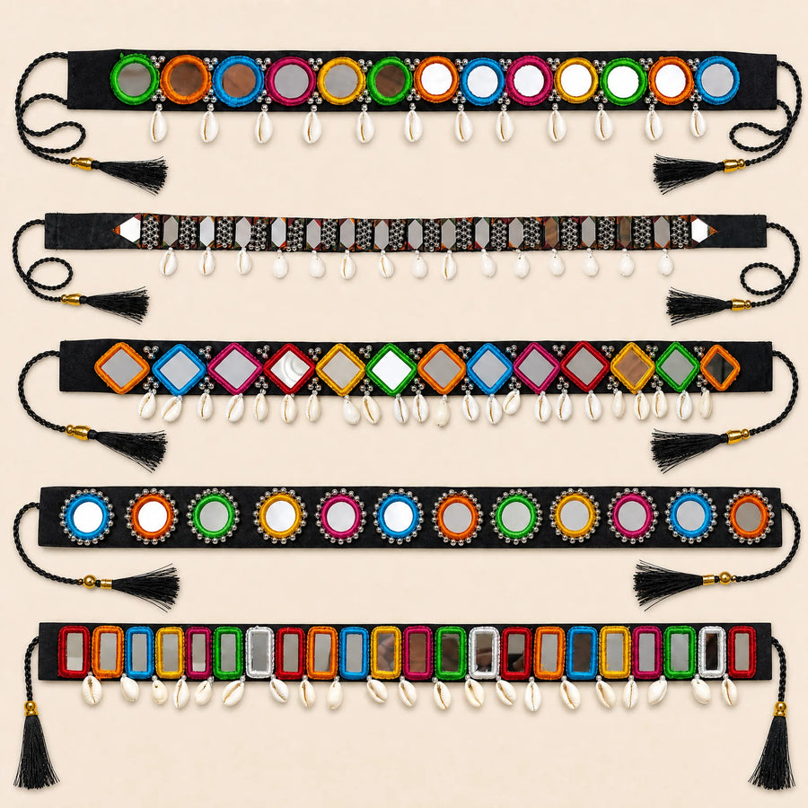 Six decorative waist belts laid flat on a light beige background. Each belt is black with colorful mirrored circular and rectangular embellishments, dangling white cowrie shells, and black tassels at both ends.